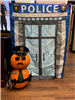 Pumpkin figure dressed as a policeman in front of a police station backdrop. Name tag reads 