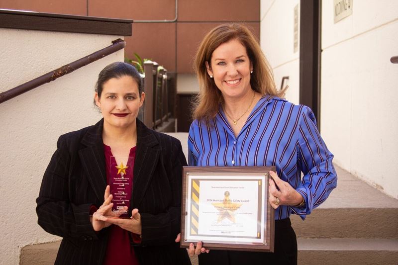 Two women stand on a staircase holding a glass trophy and a framed certificate