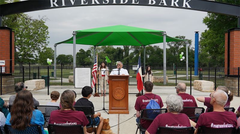 A man speaks at a podium in front of the Riverside Bark dog park. Audience sits in folding chairs