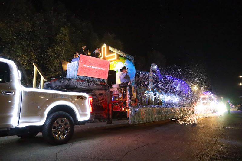 A white lighted truck pulls a lighted trailer with a snow machine, a gold lighted cross and globe
