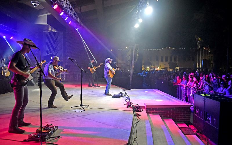 A band with multiple guitars and a fiddle performs at De Leon Plaza for a packed crowd