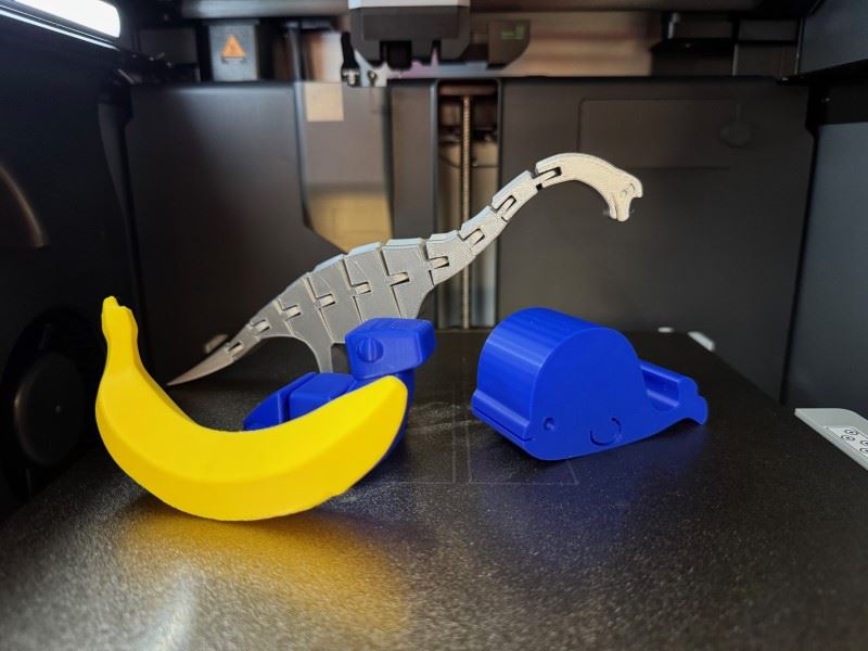 Four items created from a 3D printer including two articulated dinosaurs, a banana, and a whale.