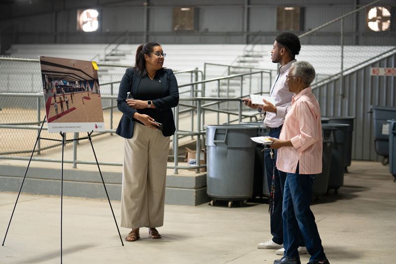 In a covered arena, a woman gestures to a posterboard with a basketball court. Two people watch.