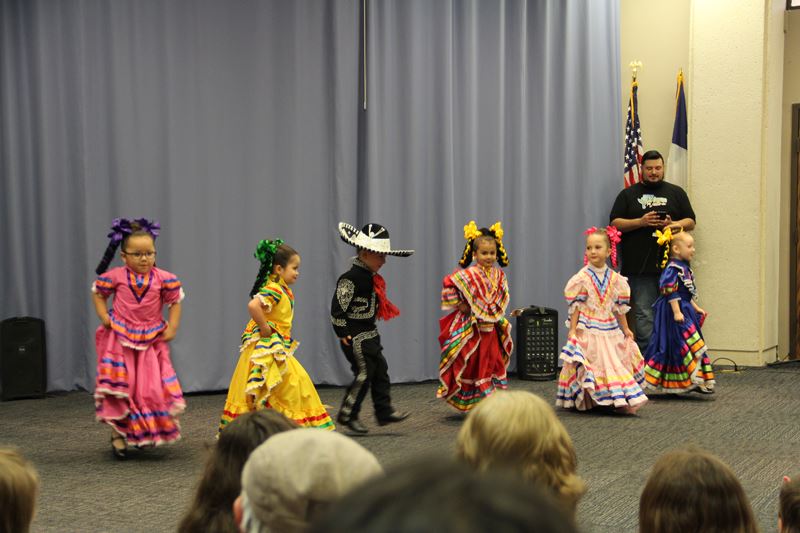 Small children in traditional Mexican dancing outfits perform indoors