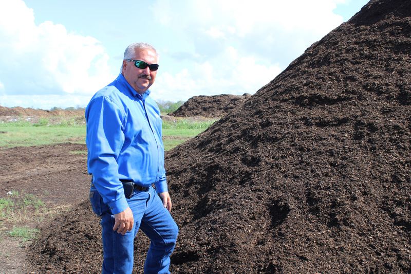 Man in a blue dress shirt and jeans stands next to a huge pile of compost