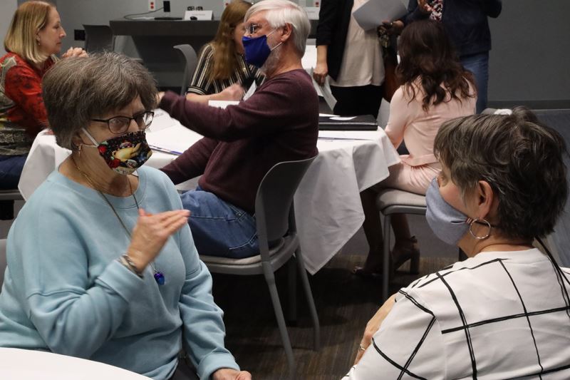 Two masked women seated at a table have a conversation. Other people are seated in the background.